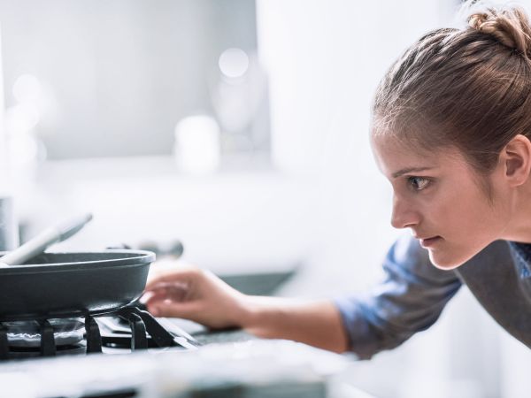 Frau steht mit eienr Pfanne an einem Gasherd und macht die Gasflamme an.
