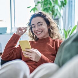 Frau sitzt mit Handy auf dem Sofa und lächelt.